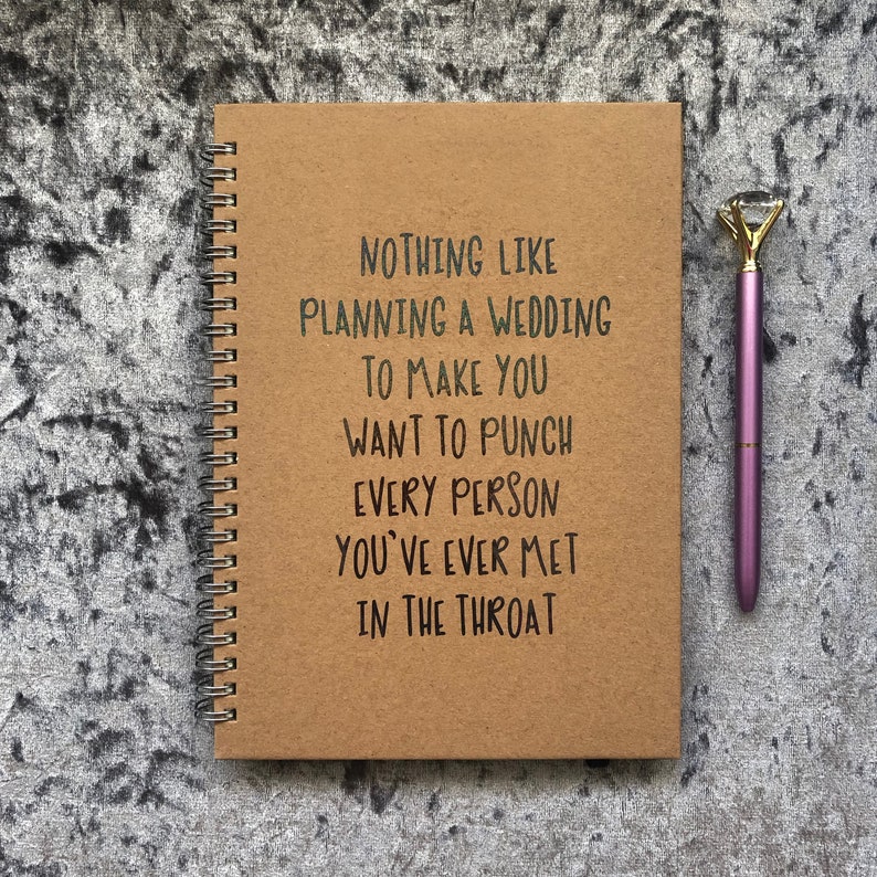 May include: A spiral-bound notebook with a brown cover and the text "NOTHING LIKE PLANNING A WEDDING TO MAKE YOU WANT TO PUNCH EVERY PERSON YOU'VE EVER MET IN THE THROAT." A purple pen with a diamond-shaped top rests beside the notebook.