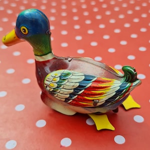 Peut inclure: Un canard en jouet vintage à remonter, avec une tête bleue, un corps brun et des ailes colorées. Le canard est en métal et possède un remontoir jaune sur le dos.