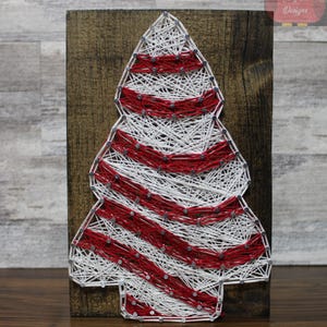 May include: A Christmas tree string art design on a wooden background. The tree is made of white and red string, with the red string forming horizontal stripes. The wooden background is a dark brown color. The design is a festive holiday decoration.