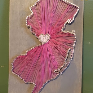 May include: Pink string art of the state of New Jersey with a white heart in the center. The art is on a gray wooden background and measures 8 inches by 14 inches. The text "8"x14" Classic Gray" is printed below the art.