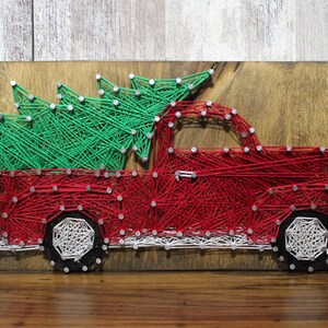 May include: String art piece featuring a red pickup truck carrying a green Christmas tree. The artwork is crafted with red and green string on a wooden board, with white accents on the wheels. The background is a natural wood grain.