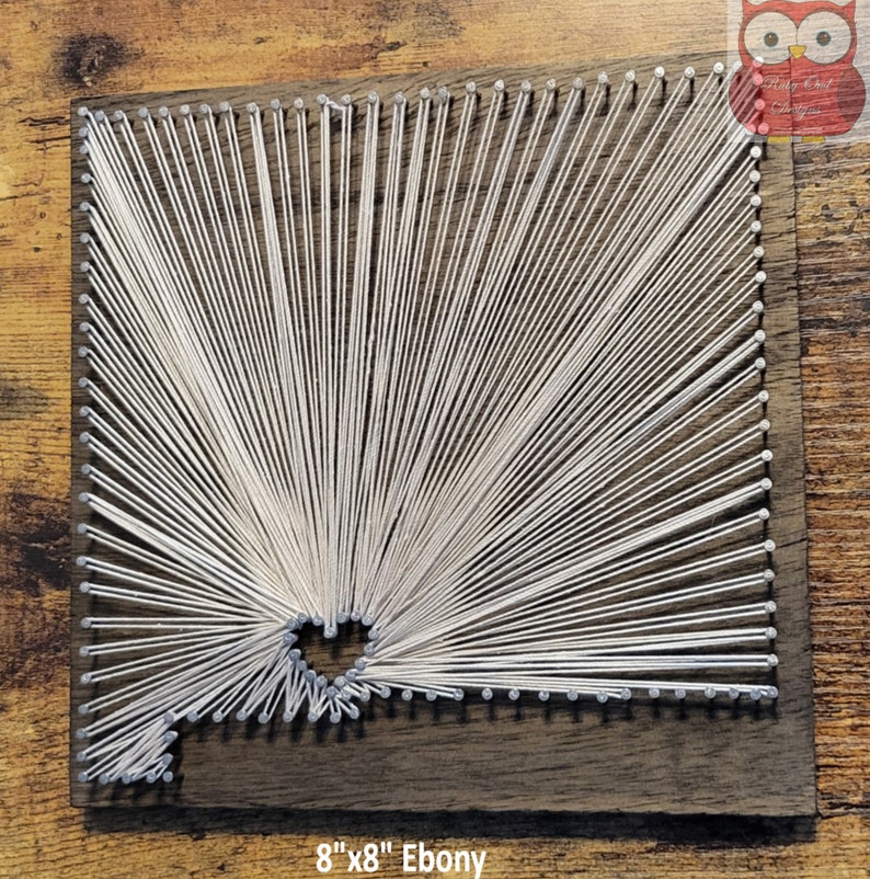 May include: A white string art design on an 8" x 8" ebony wood square. The string art design is a geometric pattern with a hole in the center.