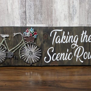 May include: Wooden sign with a string art bicycle carrying flowers in a basket. The sign is dark brown with white text that reads "Taking the Scenic Route". The bicycle is made of silver string.