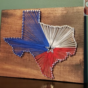 May include: A string art piece depicting the state of Texas in the shape of the state outline. The art is made with blue, white, and red string, creating the colors of the Texas flag.
