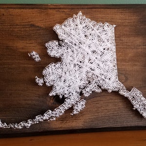 May include: White string art of the state of Alaska on a dark brown wooden background.