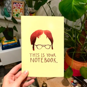 May include: A yellow notebook featuring a cartoon illustration of a person with brown hair and glasses. The text "THIS IS YOUR NOTEBOOK." is printed in brown on the front of the notebook.