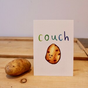 May include: A white card with the word "couch" written in rainbow colours. A cartoon potato with a smiling face is drawn below the word. A real potato is sitting on a wooden surface to the left of the card.