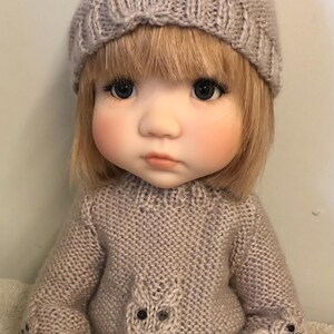 Könnte beinhalten: Puppe mit gestrickter beiger Mütze und Pullover. Der Pullover hat ein Eulenmuster auf der Vorderseite. Die Puppe hat blondes Haar und trägt eine blaue Jeans.