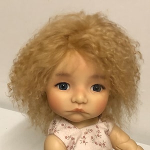 Könnte beinhalten: Eine Puppe mit blonden, lockigen Haaren, die ein rosa-weißes Blumenkleid trägt. Die Puppe hat blaue Augen und einen ernsten Gesichtsausdruck.