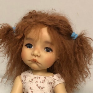 Könnte beinhalten: Eine Puppe mit langen, lockigen, rotbraunen Haaren, die zu Zöpfen mit blauen Haargummis gebunden sind. Die Puppe trägt ein weißes Kleid mit Blumenmuster und hat blaue Augen.