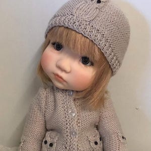 Könnte beinhalten: Eine Puppe mit einer gestrickten beigen Mütze und einem Cardigan. Die Mütze hat ein Eulenmuster mit schwarzen Knopfaugen. Der Cardigan hat Eulenmuster auf den Taschen und Ärmeln. Die Puppe hat blondes Haar und trägt eine blaue Jeans.