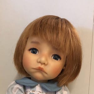 Könnte beinhalten: Eine Puppe mit einem realistischen Gesicht, blauen Augen und kurzem, rotbraunem Haar. Die Puppe hat rosige Wangen und einen leicht zusammengekniffenen Mund. Sie trägt ein blau-weiß gestreiftes Hemd mit Kragen. Der Ausdruck der Puppe wirkt nachdenklich.