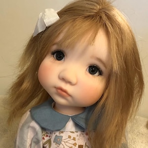 Könnte beinhalten: Handgefertigte Puppe mit blondem Haar und einer weißen Schleife. Die Puppe trägt ein blau-weißes Kleid mit floralem Muster und einem blauen Kragen. Das Gesicht der Puppe ist detailliert mit rosigen Wangen und großen, dunklen Augen.