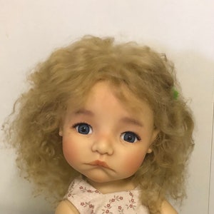 Könnte beinhalten: Nahaufnahme einer Puppe mit blonden, lockigen Haaren und blauen Augen. Die Puppe trägt ein weißes Kleid mit Blumenmuster.