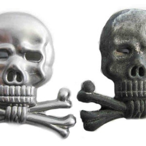 May include: Two skull and crossbones emblems. One is silver, the other is dark gray. Each skull has eye sockets, a nose cavity, and a row of teeth. The crossbones are positioned beneath the skulls.