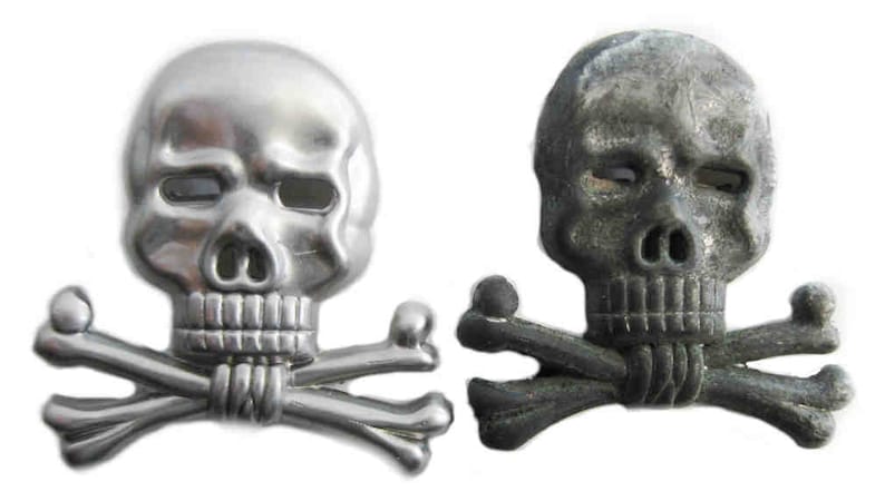 May include: Two skull and crossbones emblems. One is silver, the other is dark gray. Each skull has eye sockets, a nose cavity, and a row of teeth. The crossbones are positioned beneath the skulls.