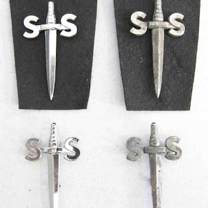 May include: Four silver-colored metal pins featuring a dagger piercing the letter "S" on each side. Two pins are mounted on black felt, while the other two are loose. The pins have a vintage or antique appearance.