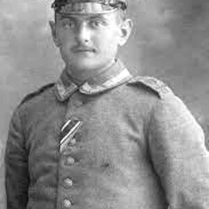 May include: Black and white portrait of a man in a military uniform. He wears a peaked cap with a badge, a button-down jacket with a medal, and a belt. The uniform has a high collar and shoulder epaulettes.