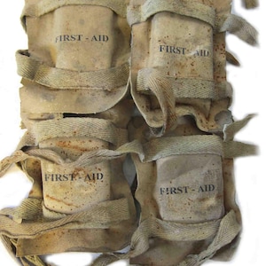 May include: A vintage first-aid kit with four compartments. Each compartment is labelled "FIRST-AID" in black text. The kit is made of aged, tan-coloured canvas and secured with woven straps.