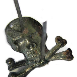 May include: A detailed, close-up view of a skull and crossbones pin. The pin is made of a tarnished, aged metal, with visible wear and discoloration. The skull has detailed features, and the crossbones are positioned beneath it. The pin's post is visible.