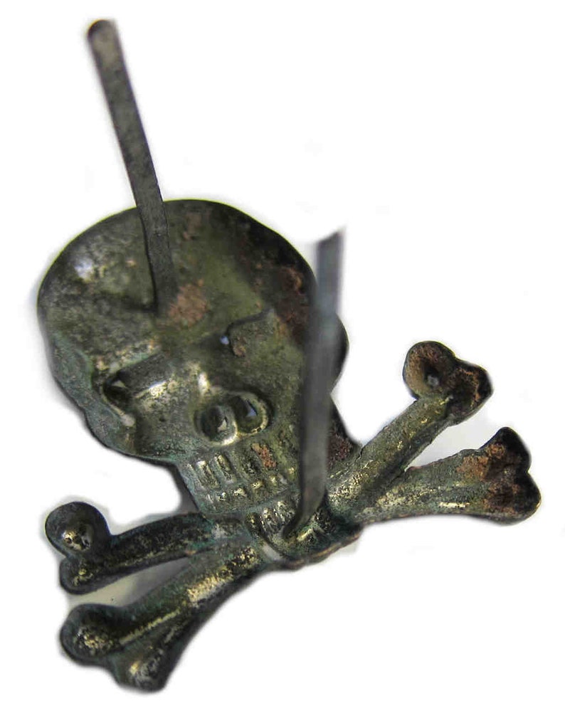 May include: A detailed, close-up view of a skull and crossbones pin. The pin is made of a tarnished, aged metal, with visible wear and discoloration. The skull has detailed features, and the crossbones are positioned beneath it. The pin's post is visible.