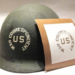 May include: A green military helmet with a white stencil that reads "WAR CORRESPONDENT US" on the side. A brown paper stencil with the same design is next to the helmet.