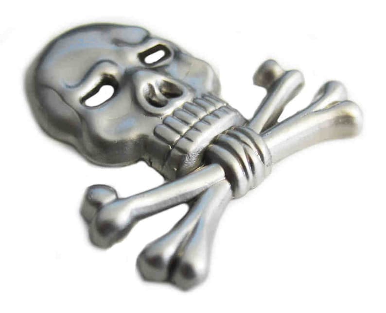 May include: A silver-colored skull and crossbones emblem. The skull has detailed features, including eye sockets, a nose cavity, and teeth. The crossbones are positioned behind the skull, creating a classic pirate symbol.
