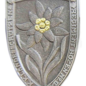 May include: A silver-toned metal badge featuring a stylised edelweiss flower in the centre. The badge is shaped like a shield and has the text "REGIMENT BOZEN" and "K.K. LANDES-SCHUTZEN" around the edge. The flower has a gold centre with five petals.