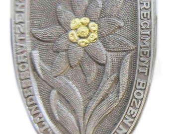 May include: A silver-toned metal badge with a stylized edelweiss flower in the center. The badge is shaped like a shield and has the text "REGIMENT BOZEN" and "K.K. LANDES-SCHUTZEN" around the edge. The flower has a gold center with five petals.