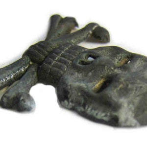May include: A small, antique-style metal skull and crossbones charm. The charm is a dark, tarnished gray color with visible details. The skull has eye sockets and the crossbones are tied together. This item is likely a decorative element or jewelry component.