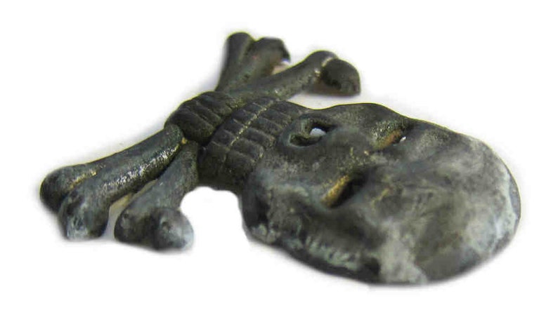 May include: A small, antique-style metal skull and crossbones charm. The charm is a dark, tarnished gray color with visible details. The skull has eye sockets and the crossbones are tied together. This item is likely a decorative element or jewelry component.