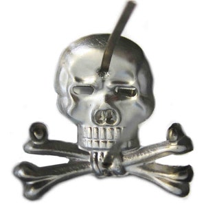 May include: A silver-colored skull and crossbones emblem with a metal rod piercing the skull. The skull has detailed features, and the crossbones are positioned beneath it. The emblem is likely a decorative pin or badge.