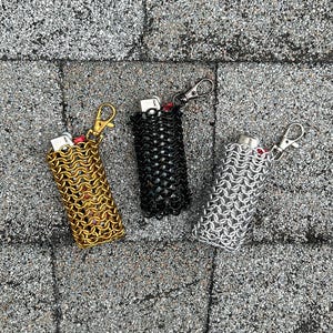Könnte beinhalten: Drei Feuerzeug-Etuis im Kettenstil in Gold, Schwarz und Silber. Jedes Etui hat einen passenden Metallverschluss und einen roten Feuerzeugknopf. Die Feuerzeuge liegen auf einer strukturierten grauen Oberfläche.