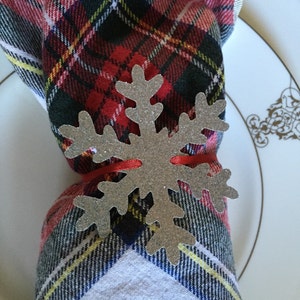 Op de afbeelding: Een zilveren sneeuwvlok servetring met een rood lint eromheen gebonden. De servetring ligt op een rood-zwarte geruite servet.