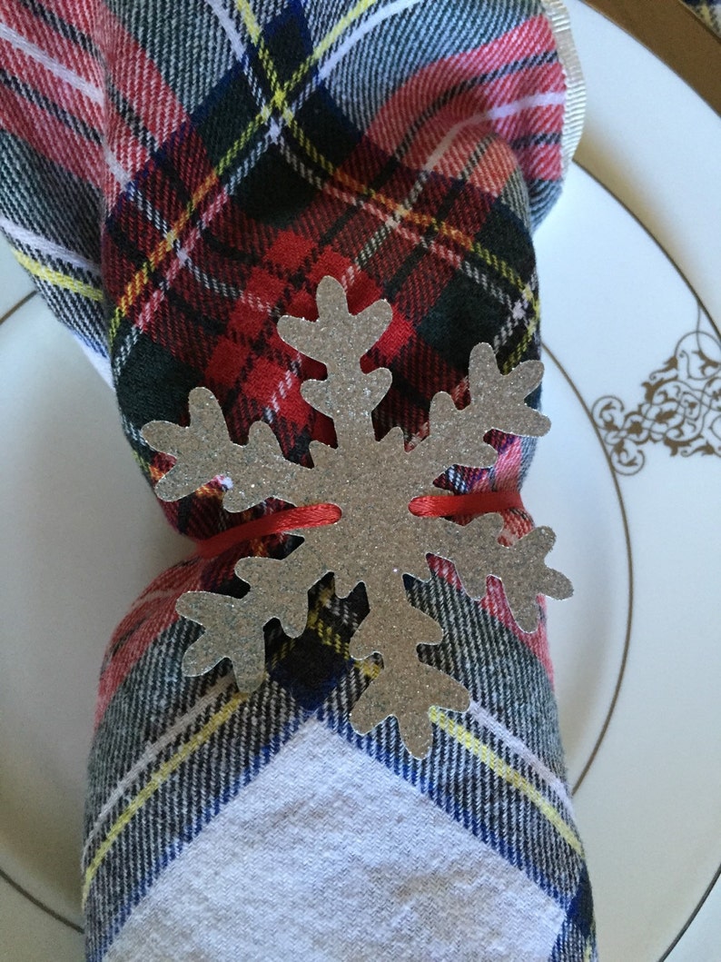 Op de afbeelding: Een zilveren sneeuwvlok servetring met een rood lint eromheen gebonden. De servetring ligt op een rood-zwarte geruite servet.