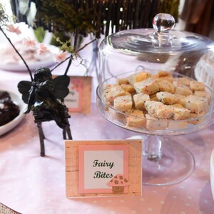 May include: A glass cloche with a white and pink sign that reads "Fairy Bites" sits on a table with a pink tablecloth. The cloche is filled with white sugar cookies with colorful sprinkles. There is a small black fairy figurine on the table.