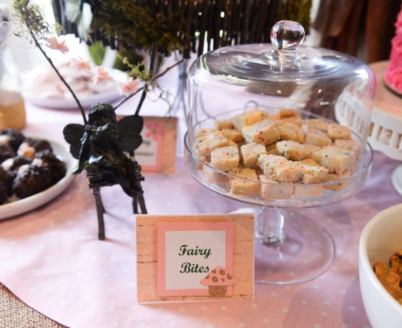 May include: A glass cloche with a white and pink sign that reads "Fairy Bites" sits on a table with a pink tablecloth. The cloche is filled with white sugar cookies with colorful sprinkles. There is a small black fairy figurine on the table.