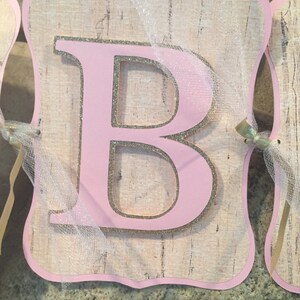 May include: Pink letter "B" with gold glitter accents on a woodgrain background. The letter is attached to a white tulle ribbon.