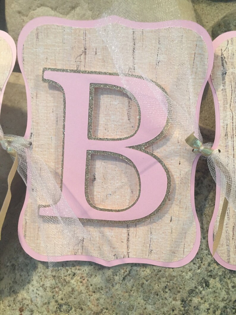 May include: Pink letter "B" with gold glitter accents on a woodgrain background. The letter is attached to a white tulle ribbon.