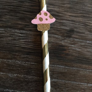 May include: A gold and white striped paper straw with a pink and gold glitter mushroom attached to the top.