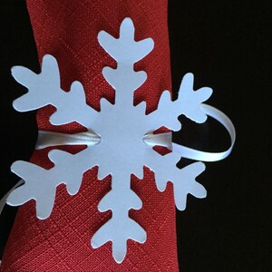 Op de afbeelding: Witte papieren sneeuwvlok servetring op een rode servet. De sneeuwvlok heeft een eenvoudig ontwerp met zes punten.
