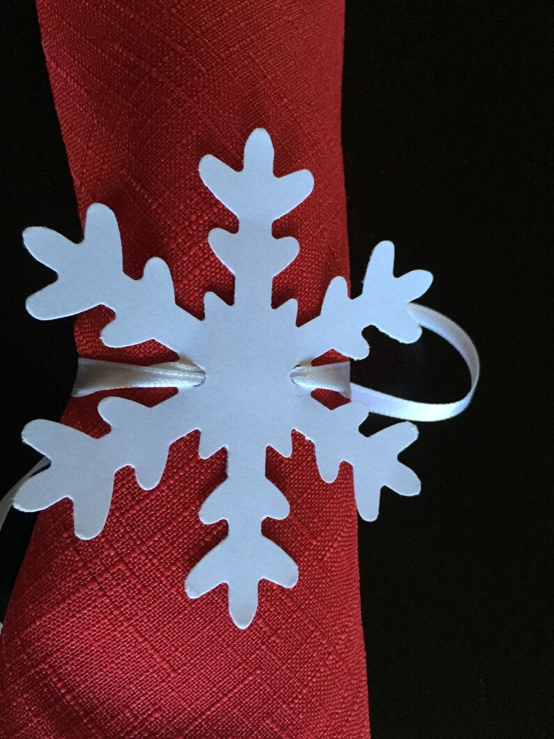 Op de afbeelding: Witte papieren sneeuwvlok servetring op een rode servet. De sneeuwvlok heeft een eenvoudig ontwerp met zes punten.