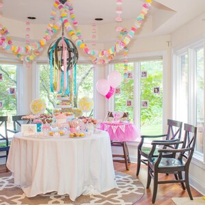 May include: A white table with a white tablecloth is set for a party. The table is decorated with pink and blue balloons, and a variety of desserts. There are also two chairs with black frames and white cushions.