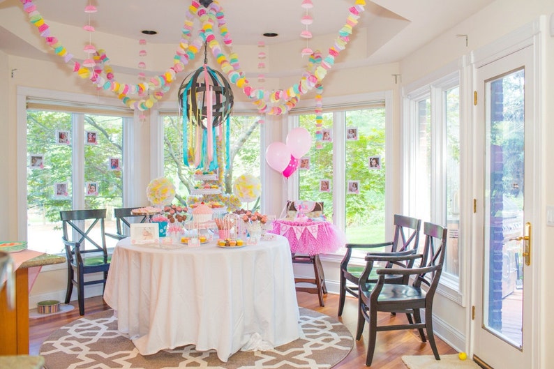 May include: A white table with a white tablecloth is set for a party. The table is decorated with pink and blue balloons, and a variety of desserts. There are also two chairs with black frames and white cushions.
