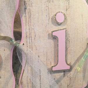 May include: Pink and gold letter "I" with glitter accents on a beige background. The letter is part of a banner with a white tulle ribbon.