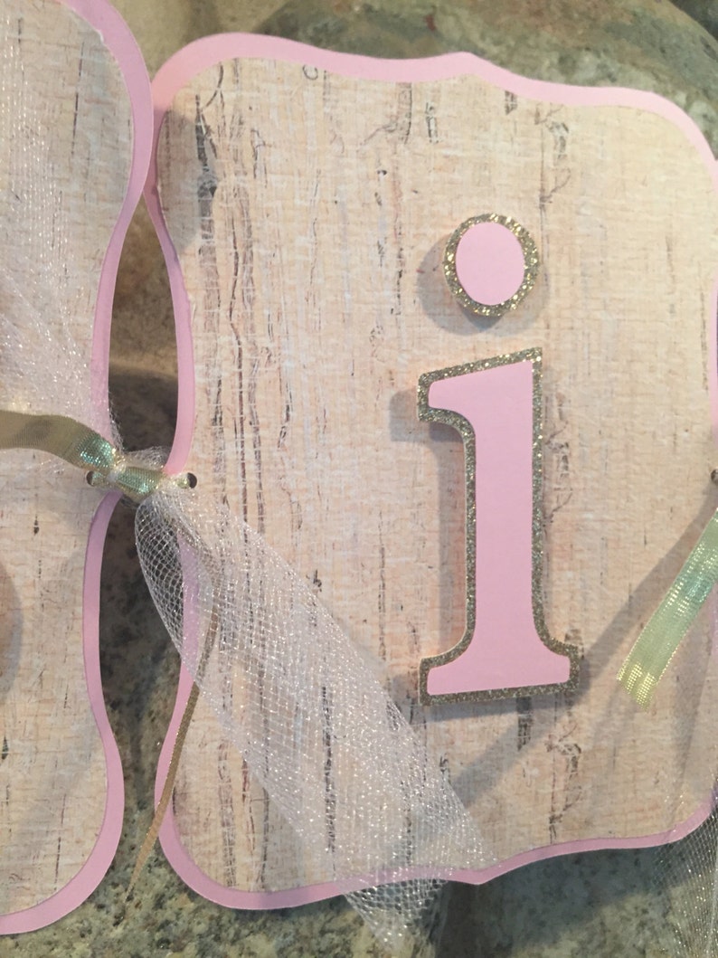 May include: Pink and gold letter "I" with glitter accents on a beige background. The letter is part of a banner with a white tulle ribbon.