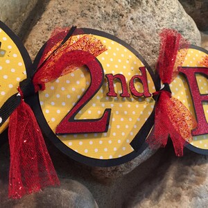 Puede incluir: Una pancarta amarilla y negra con letras rojas de purpurina que dicen "2nd Birthday". La pancarta tiene lunares blancos y lazos de cinta roja y negra.