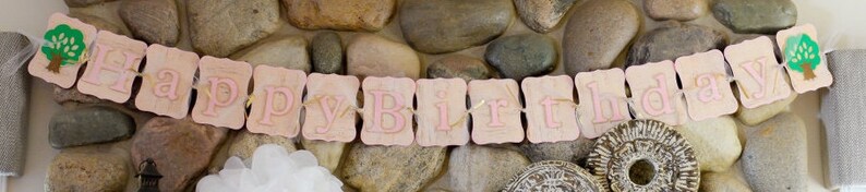 May include: A pink and white banner with the words "Happy Birthday" in pink letters. The banner is decorated with small green trees on each end.