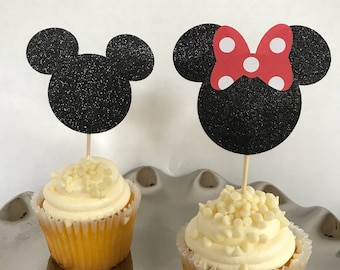 Könnte beinhalten: Zwei Cupcakes mit weißem Zuckerguss und weißen Schokoladenstückchen. Einer hat einen schwarzen Glitzer-Mickey-Mouse-Kopf-Topper, der andere einen Minnie-Mouse-Kopf-Topper mit einer roten Schleife mit weißen Punkten. Die Cupcakes stehen auf einem silbernen Tablett.