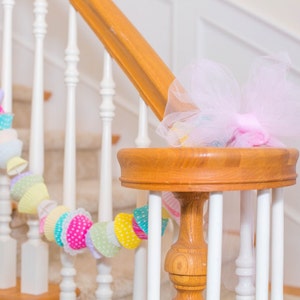 May include: A white staircase with a wooden handrail decorated with a colorful garland of paper cupcake liners and a pink tulle bow.